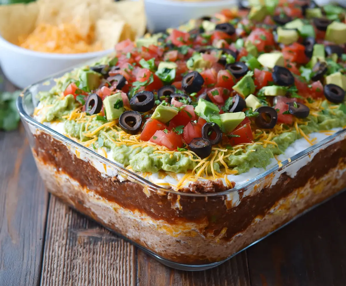 Delicious Ultimate 7 Layer Dip featuring guacamole, sour cream, shredded cheese, tomatoes, olives, and green onions served in a clear glass dish.